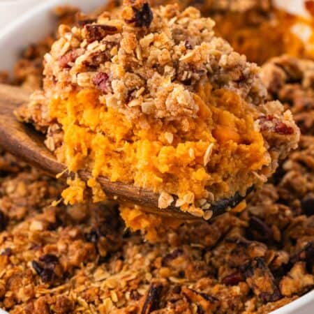 A wooden spoon lifting a scoop of mashed sweet potato filling and pecan streusel out of the baking dish.
