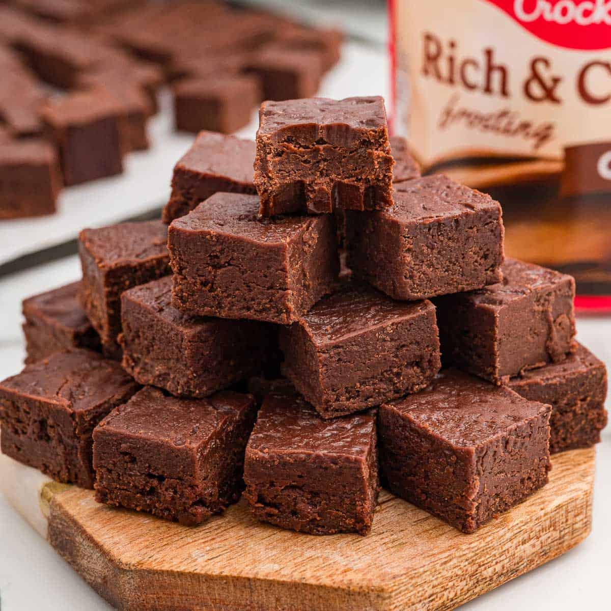 Fudge squares stacked on a wooden board with the top piece missing a bite and a frosting tub in the background.