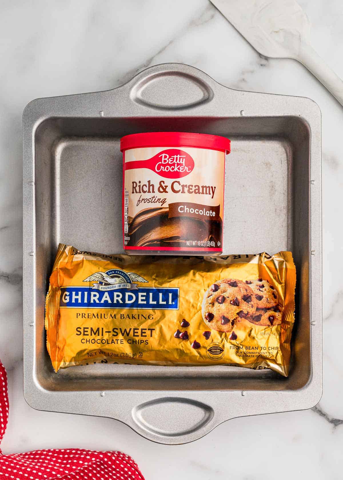 Ingredients in a square metal pan: a tub of Betty Crocker Rich & Creamy chocolate frosting and a bag of Ghirardelli semi-sweet chocolate chips.