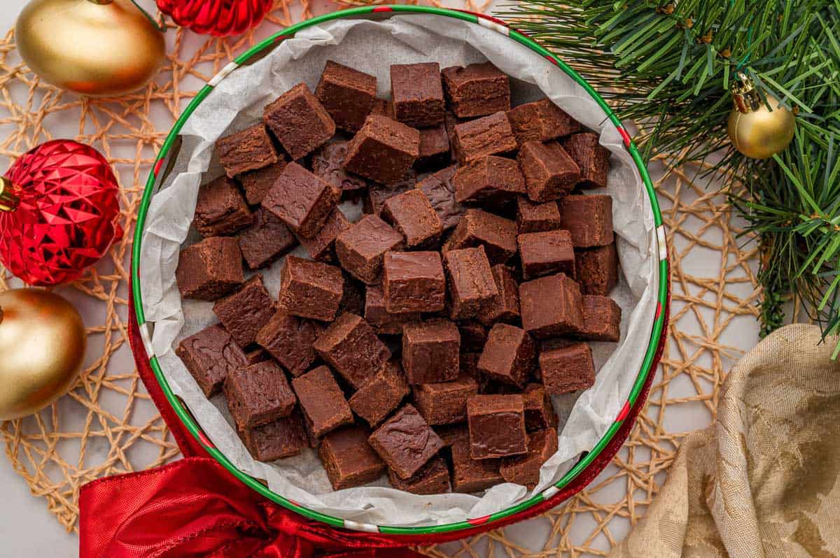 Squares of 2 ingredient fudge with frosting piled in a round holiday tin lined with parchment paper, with red and gold ornaments and pine branches around it.