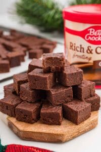 Pyramid of chocolate fudge squares stacked on a round wooden board with a tub of chocolate frosting behind it.