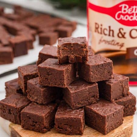 Pyramid of chocolate fudge squares stacked on a round wooden board with a tub of chocolate frosting behind it.