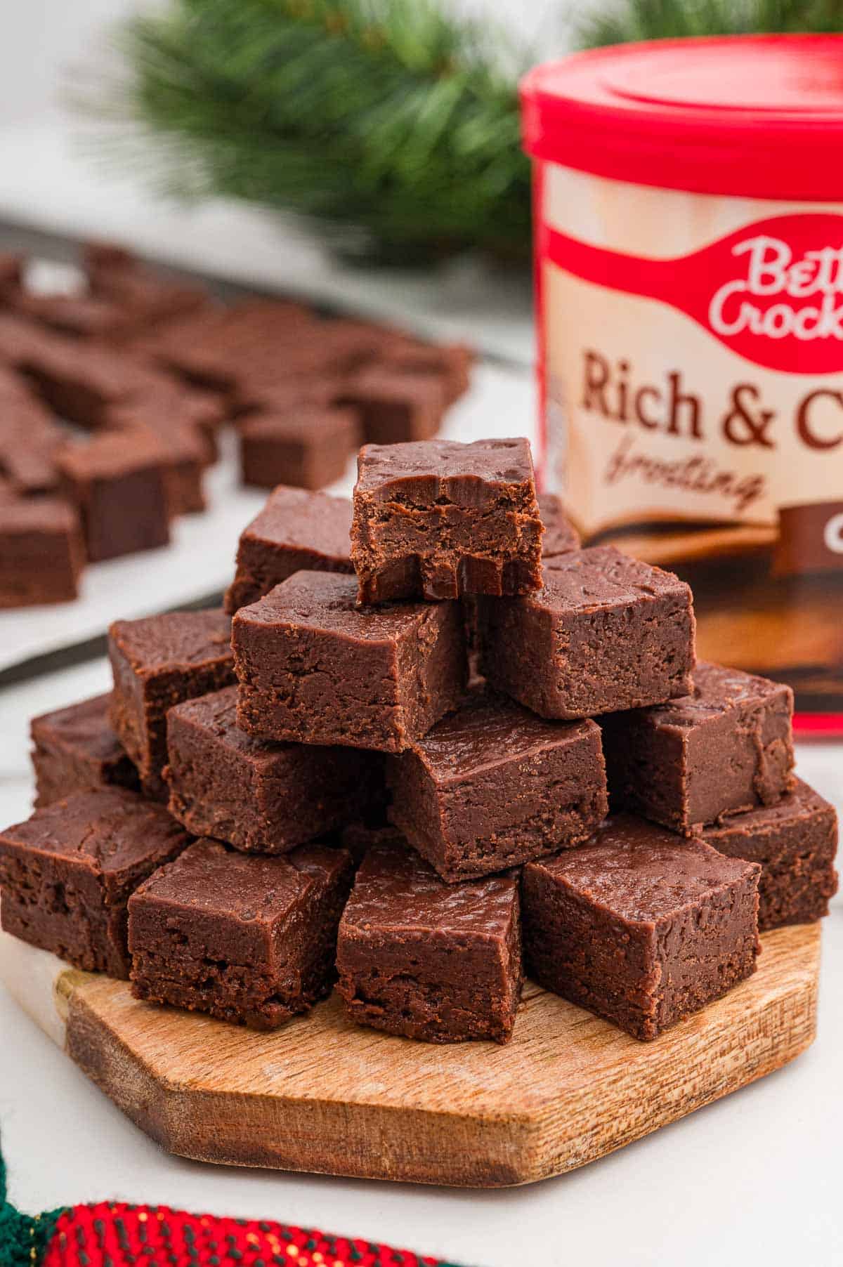 Pyramid of chocolate fudge squares stacked on a round wooden board with a tub of chocolate frosting behind it.