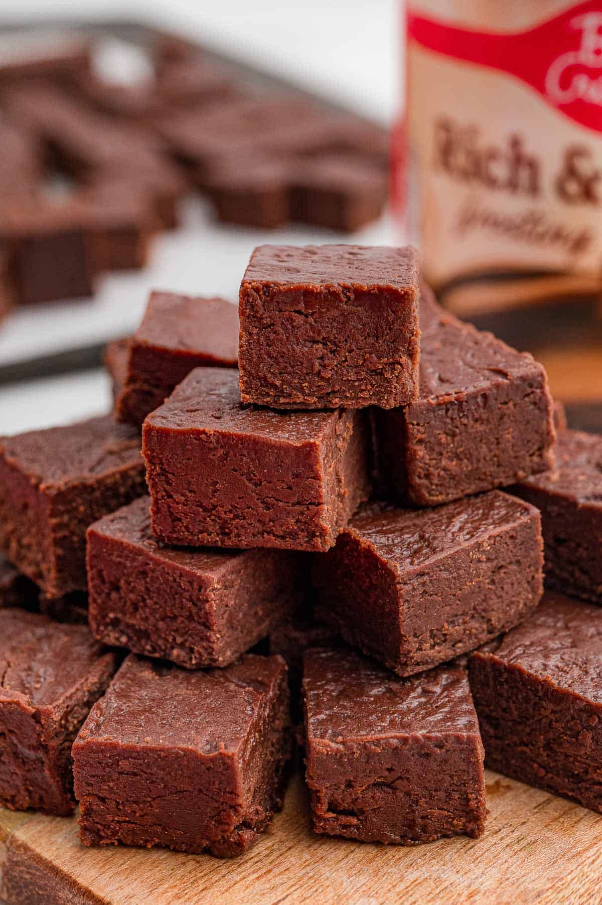 Close-up of chocolate fudge squares stacked on a wooden board with a frosting tub behind them.