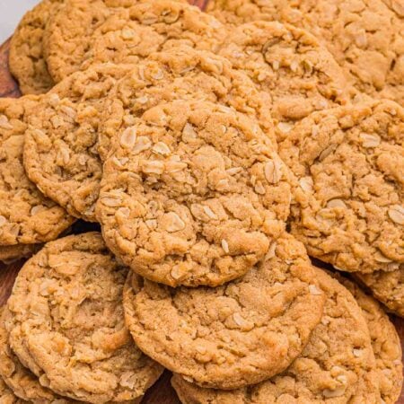Cookies stacked and overlapping on a wooden board, with a jar of oats and a bowl of peanut butter behind.