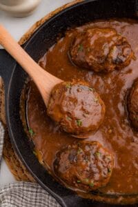 A wooden spoon lifting a gravy-covered beef patty out of a cast iron skillet.