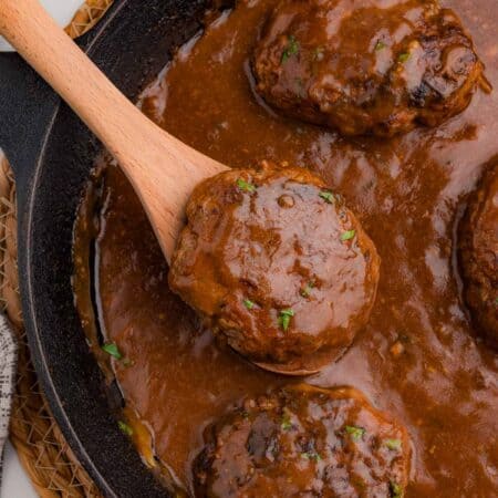 A wooden spoon lifting a gravy-covered beef patty out of a cast iron skillet.