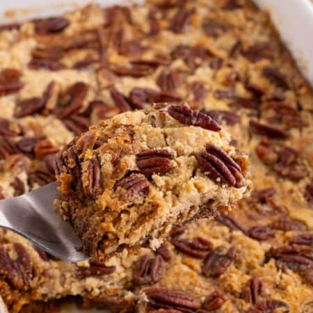 A spatula lifting a piece of cake from a white baking dish, with gooey pecan filling left in the pan.